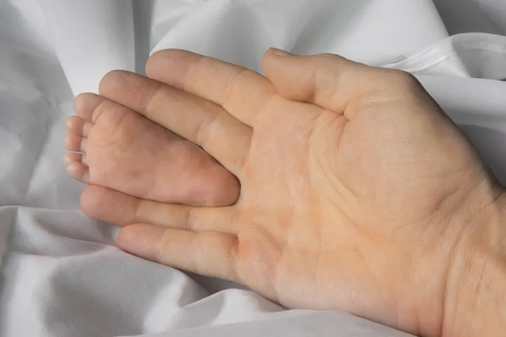Sole of foot of an infant held between the middle finger and the ring finger of an adult hand for size comparison